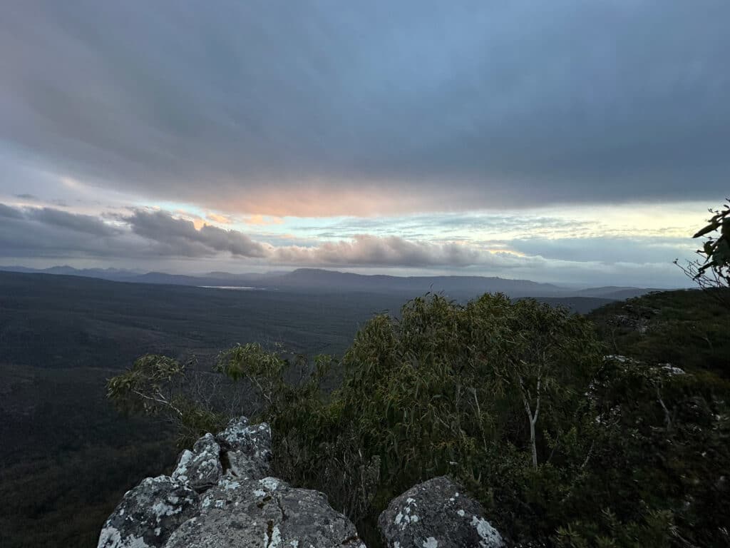 A panoramic view of a vast landscape with distant mountains, rocky outcrops, and dense foliage during sunrise or sunset, with the sky painted in orange, pink, and blue hues